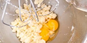 A mixing bowl with two cracked eggs and dense, crumbly ingredients is positioned under metal beater attachments. A liquid is being poured into the mixture from a measuring cup on the right side of the image.