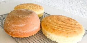 Three round loaves of bread are cooling on a wire rack. The loaf on the left is slightly darker and appears to have a thicker crust, while the other two loaves are lighter in color with visible horizontal lines on their surfaces. The background shows a white tile wall.