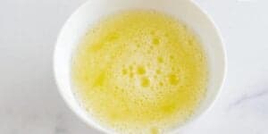 A white bowl filled with frothy beaten egg whites, featuring a light yellow color. The bowl is placed on a white surface.
