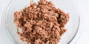 A glass bowl filled with a mixture of raw ground meat and finely chopped onions. The meat appears to be seasoned with various spices and herbs. The bowl is placed on a light-colored surface.