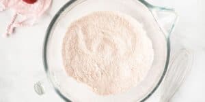 A clear glass mixing bowl filled with a light pink powder sits on a white surface. A metal whisk is placed beside the bowl, and part of a pink cloth is visible in the corner of the image.