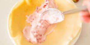 A close-up image of a spoon spreading a creamy pink mixture with visible strawberry chunks into an unbaked pie crust. The unbaked pie crust is in a white pie dish.