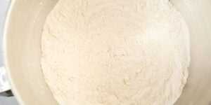 A large metal mixing bowl filled with finely milled white flour. The flour is spread evenly across the bottom of the bowl, creating a smooth, powdery surface. The edges of the bowl are slightly visible.