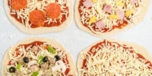 Four homemade pizzas with different toppings are shown. One pizza has pepperoni, another ham and pineapple, a third features mushrooms, olives, green peppers, and sausage, and the last one is just cheese. The pizzas are on a light-colored surface.