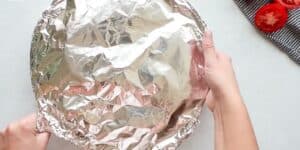 Hands are holding a round baking dish covered with aluminum foil. There is a striped cloth with two sliced tomatoes near the top right corner of the image. The background is a clean, light-colored surface.