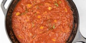 A close-up view of a cast iron skillet filled with rich, red tomato sauce. The sauce appears chunky with visible pieces of vegetables and herbs mixed in, suggesting a homemade or rustic style of cooking.