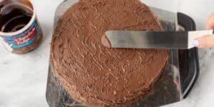A round chocolate cake being frosted with chocolate fudge icing. A hand holding an offset spatula is seen smoothing the frosting on the top of the cake. A tub of chocolate fudge icing is in the background on a marble surface.