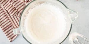 A glass mixing bowl containing whipped cream is shown from above. A whisk with some whipped cream on it and a red and white striped kitchen towel are placed next to the bowl on a light gray countertop.
