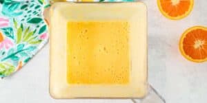 A top-down view of a blender filled with a creamy orange-colored liquid. Beside the blender, there are two halves of an orange with a colorful floral-patterned cloth on the left side of the image.