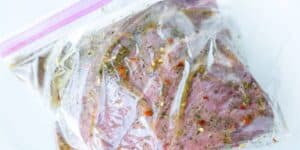 A plastic zip-top bag filled with marinated meat. The marinade contains herbs, spices, and small red chili flakes, coating the raw meat evenly. The bag is sealed and placed on a white surface.