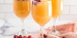 Three wine glasses filled with a yellow-orange fruit punch garnished with cranberries. The glasses are placed on a white marble surface, with a pink cloth and scattered cranberries beside them. The background consists of a white tiled wall.
