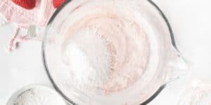A top view of a mixing bowl with a pink batter and white powder, possibly confectioner's sugar, being added. Nearby are a whisk, a bowl with more white powder, and a pink cloth with three strawberries on it. The scene is set on a white surface.