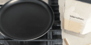 Close-up photo of a stove burner with a black pan on the left side and a Blendtec blender container filled with light-colored contents, marked with measurement lines, on the right, standing on a white surface.