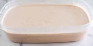 A rectangular plastic container filled with light brown ice cream rests on a white surface. The smooth surface of the ice cream indicates it has been freshly made or recently stored. The container's lid is not shown in the image.