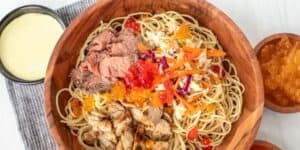 A wooden bowl filled with noodles, sliced beef, colorful vegetables, and fish roe sits on a white table. A small wooden bowl of orange fish roe and a small black bowl of creamy sauce are also present. The bowl is placed on a striped gray napkin.