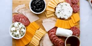 A wooden charcuterie board displaying an assortment of sliced meats, cheeses, and crackers. There are bowls containing black olives and marinated mozzarella balls. A selection of cheddar, gouda, and soft cheeses is artfully arranged alongside the meats.
