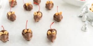 A tray of chocolate-covered cherries arranged to resemble mice, with each mouse featuring almond slices for ears and a red candy string for a tail. Silver-wrapped chocolate candies and a bowl of almonds are in the background.