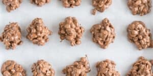 Several clusters of chocolate-covered peanuts are arranged on a white parchment paper. The clusters have an irregular, chunky shape, each appearing textured and glossy from the melted chocolate coating. The treats are evenly spaced in neat rows.