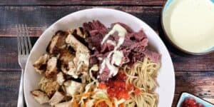 A white bowl containing grilled chicken, slices of roast beef with white sauce, noodles, and assorted vegetables garnished with red fish roe. Next to the bowl is a cup of creamy yellow soup and a small dish of dipping sauce, with a fork placed to the left of the bowl.