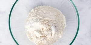 A glass bowl filled with white flour sits on a white marble surface. The flour appears finely milled and is slightly heaped towards the center of the bowl.