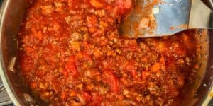 A close-up view of a pot filled with chunky, red tomato-based meat sauce. Visible ingredients include ground meat, chopped tomatoes, and bits of other vegetables. A metal spoon is partially submerged in the sauce, resting on the pot's edge.