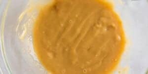 A close-up view of a bowl containing thick, caramel-colored batter or sauce. The mixture has a smooth texture with some small lumps and air bubbles visible on the surface. The bowl is transparent, allowing visibility of the mixture's consistency.