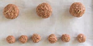 A baking sheet lined with parchment paper displaying ten uncooked meatballs. Three larger meatballs are positioned in the top row, while seven smaller meatballs are arranged evenly in the bottom row. The meat mixture is visibly seasoned.