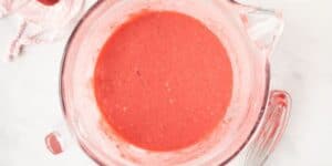 A glass mixing bowl sits on a white surface, containing a vibrant pink batter. A metal whisk lies next to the bowl, indicating that the contents have been recently mixed. The background features a faint glimpse of a pink cloth and scattered pink powder.
