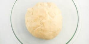 A ball of uncooked dough rests in a transparent glass bowl on a white surface. The dough is light in color and has a slightly rough texture with visible creases.