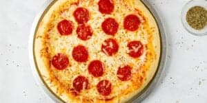 A pepperoni pizza on a round metal baking tray is displayed on a white surface. The pizza has a golden crust, melted cheese, and evenly spaced pepperoni slices. To the right of the tray, there is a small clear dish containing dried herbs.