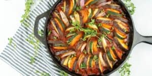 A cast iron skillet filled with a beautifully arranged ratatouille, featuring layers of zucchini, eggplant, and tomato slices. Fresh thyme garnishes the dish, and the skillet sits on a striped cloth with sprigs of thyme scattered around.