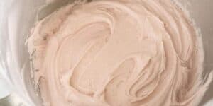 A close-up shot of a stainless steel bowl filled with creamy, light pink frosting. The frosting has a smooth, fluffy texture and has been swirled evenly around the bowl, indicating it's been freshly mixed.