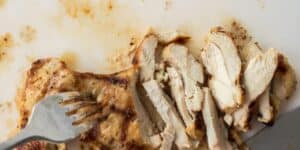A piece of grilled chicken breast is partially sliced on a white cutting board. A metal fork rests on the left side, holding down the chicken. Juices from the chicken are visible on the cutting board. The chicken appears to be seasoned and cooked to a golden brown.