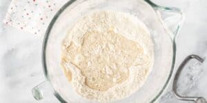 A glass mixing bowl with flour and a pool of liquid in the center, likely the beginning stages of making dough. Next to the bowl is a white cloth with a small red and blue dotted pattern. The setup is on a white marble surface.