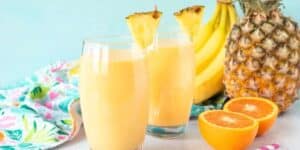 Two glasses of yellow smoothie are garnished with pineapple slices. In the background, there are whole bananas, a whole pineapple, and a halved orange. A colorful floral cloth is draped on the surface near the glasses. The background is light blue.