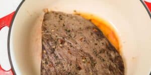 A seasoned brisket searing in a red and white Dutch oven. The meat is browned with visible herbs and spices, and there is a small amount of liquid at the bottom of the pot.