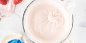 A clear mixing bowl filled with light pink whipped frosting sits on a marble surface. Part of a container with a blue label and a textured pink cloth are visible near the bowl, along with some fresh strawberries.