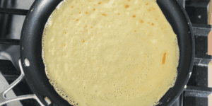 A close-up view of a crepe cooking in a black non-stick frying pan. The crepe is lightly browned with small bubbles forming on the surface. The pan is on a black stove with metallic handles visible on the left side.