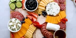 A charcuterie board with an assortment of sliced cheeses, cured meats, and fresh fruits and vegetables, including strawberries, blueberries, cucumber slices, and a cup of mozzarella balls. There are dips, spreads, and a block of goat cheese also on the board.