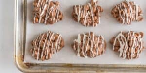 Six clusters of milk chocolate and peanuts drizzled with white chocolate are on a parchment-lined baking sheet.