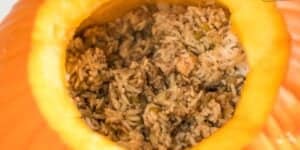 A close-up of a halved pumpkin filled with a savory mixture of cooked rice, ground meat, and diced vegetables. The blend of ingredients inside the pumpkin appears to be a hearty, autumn-inspired dish.