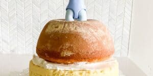 A two-tiered cake is topped with a doughnut-like pastry. An unexpected decoration, a small blue toy figure, is partially embedded in the top layer, adding a humorous touch. The background features a white, leaf-patterned wallpaper.