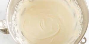 A close-up of a mixing bowl filled with smooth, creamy batter. The bowl has a metallic finish and the light-colored batter appears to have been recently mixed, with swirls still visible on its surface. The background is a light-colored countertop.