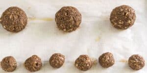 Seven small and three large raw vegan meatballs are evenly spaced on a parchment-lined baking sheet, ready to be baked. The meatballs have a textured appearance, suggesting ingredients like nuts, seeds, and vegetables.
