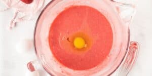 A glass mixing bowl filled with pink cake batter topped with a cracked raw egg. The bowl is on a white surface next to two whole eggs and a whisk. A pink cloth with a strawberry on it is visible in the upper left corner.