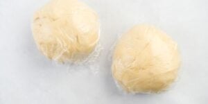 Two round balls of dough wrapped in transparent plastic wrap are placed on a white surface. The dough appears smooth and light beige in color.