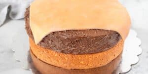 A partially covered cake with a plain, lightly browned layer of rolled fondant being applied to the top. The cake has a golden-brown base and is frosted with chocolate icing. It sits on a reflective, scalloped-edge cake board.