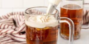 A clear glass mug filled with a brown, fizzy drink is being topped with whipped cream from a piping bag. Another filled mug is in the background, with a striped red and white cloth napkin nearby. The setting appears to be a kitchen with white tiled walls.