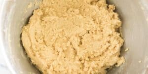 A large mixing bowl filled with thick, creamy cookie dough. The dough appears well-mixed and has a smooth texture, featuring a light tan color with small flecks throughout. The bowl is metallic and placed on a marbled surface.