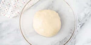 A ball of dough rests in a clear glass bowl on a white marble surface. A white cloth with small multicolored dots is partially visible in the top left corner.